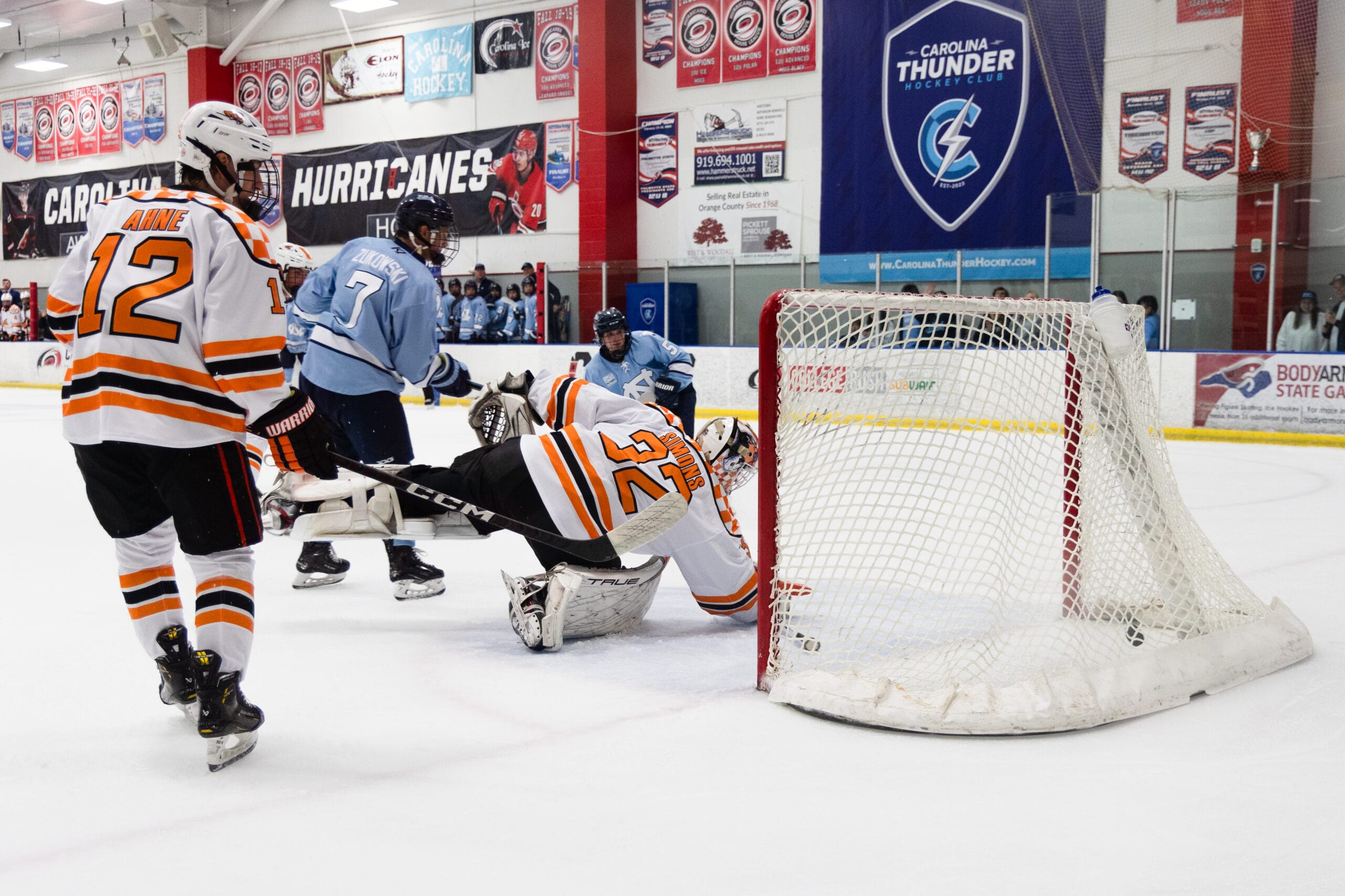 A UNC Hockey player in a light blue jersey scores past a diving University of Tennessee goalie in an orange and white uniform during a game, as another Tennessee player skates nearby.