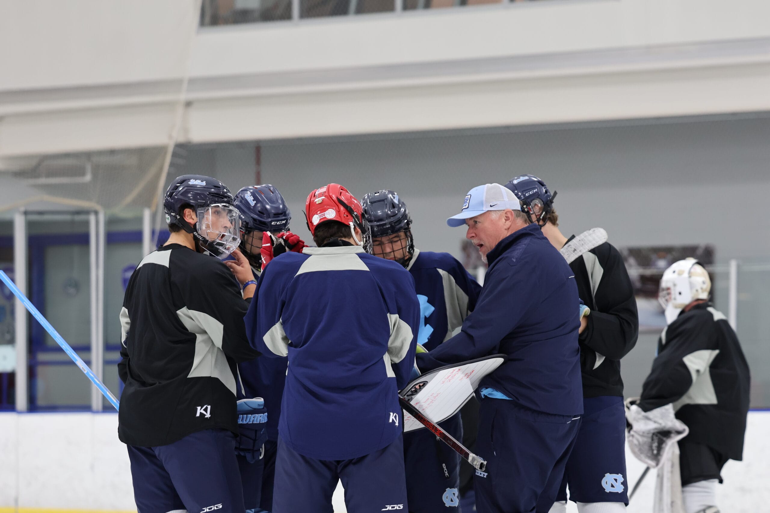 UNC Hockey head coach Steve Rice, holding a dry erase board, speaks to players gathered around him on the ice during practice.